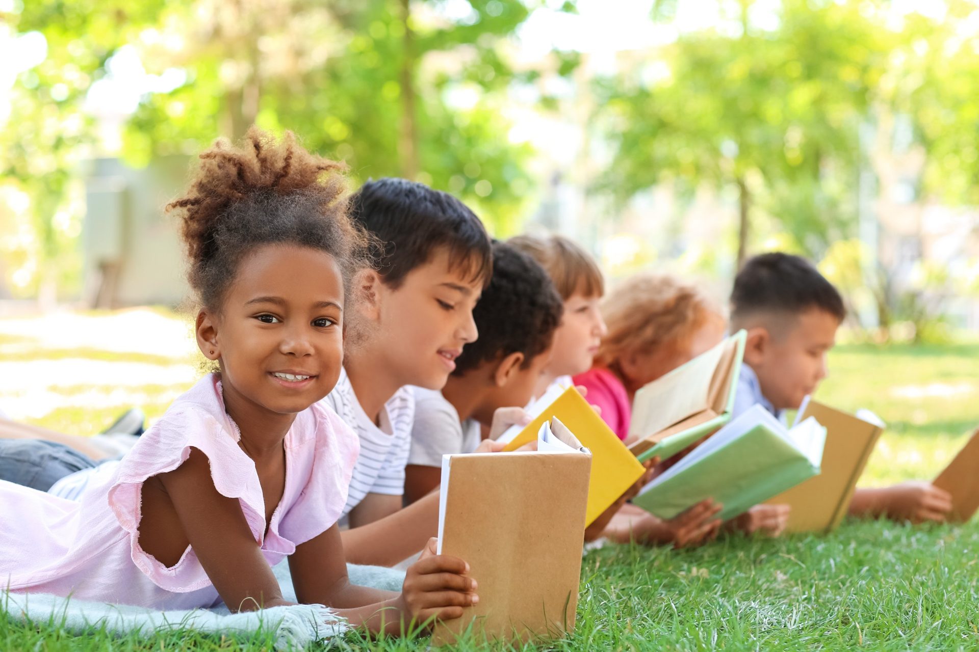 A smiling girl lies on the grass with a book, beside a row of children reading together in the sunshine