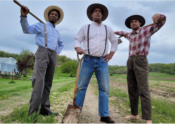 Three performers in period work clothes and hats stand on a farm path holding a shovel and hoes